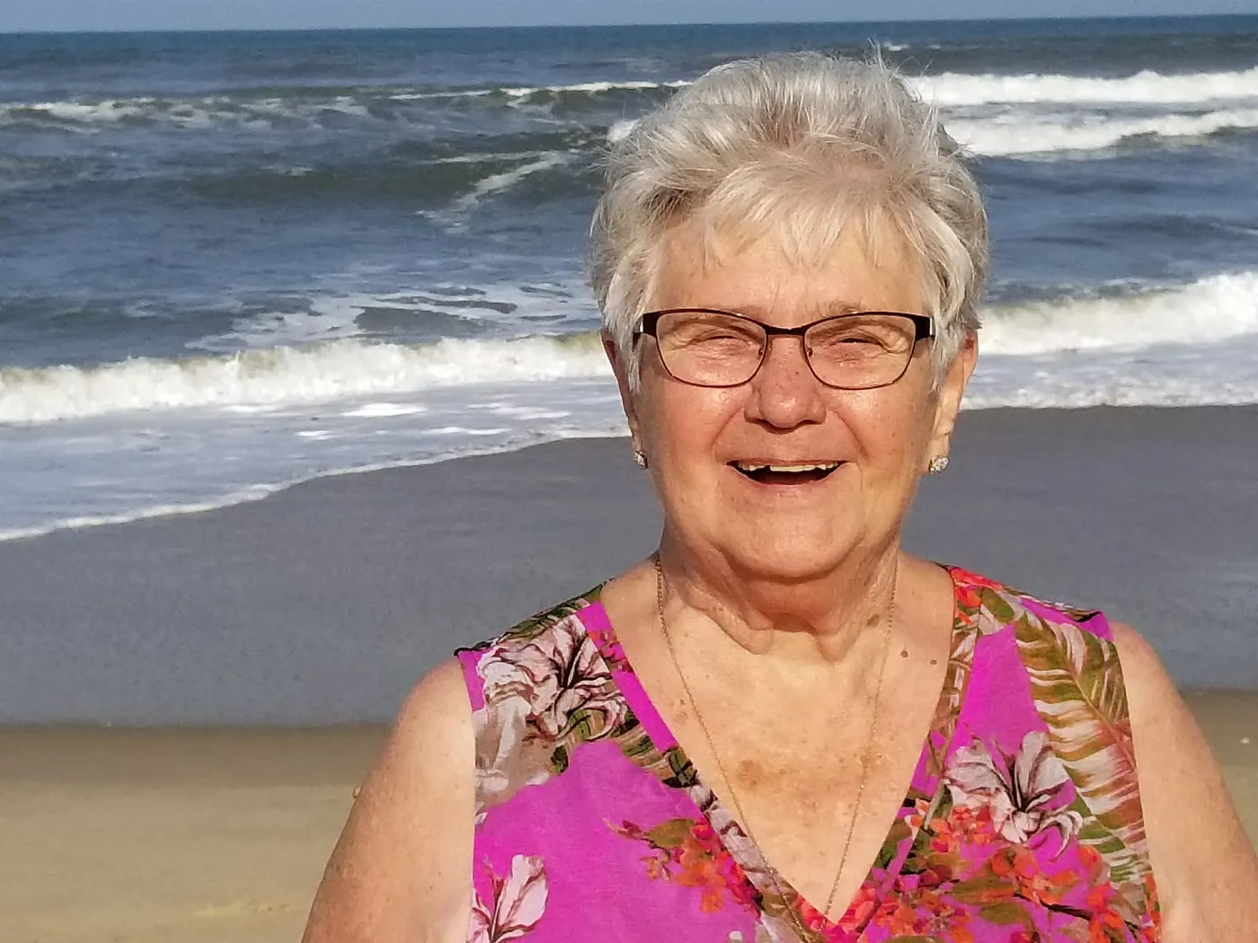 Dolores laughing on the beach in a bright pink floral top, white-capped waves rolling in behind her.