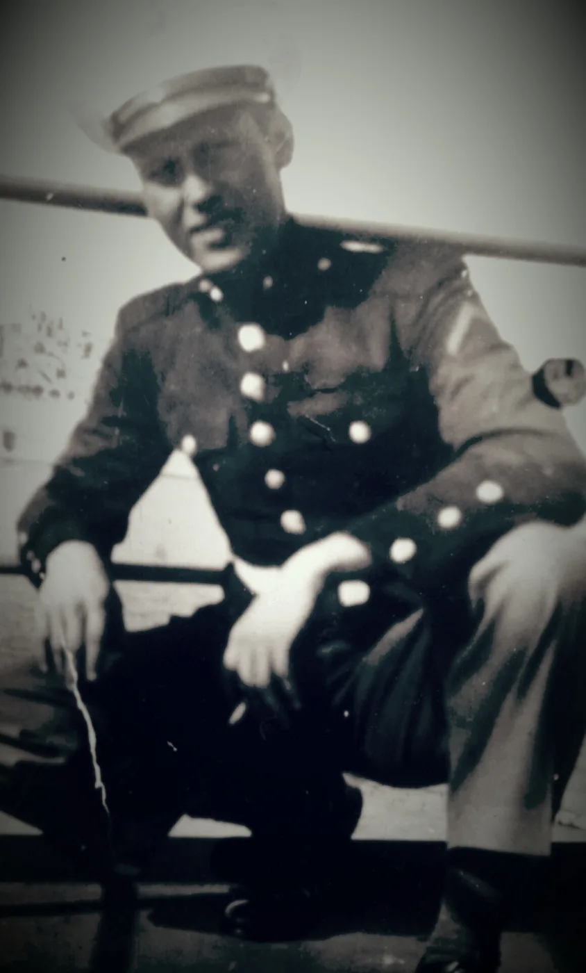 Faded black-and-white photograph of a young man in Marine Corps dress blues and a white cap, crouching beside a rail with his hands resting on his knees.