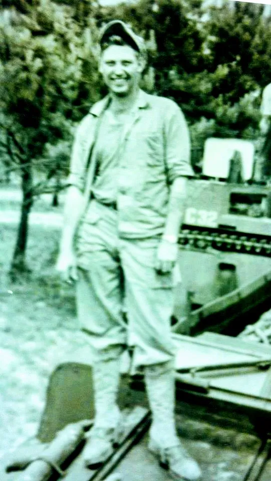 Black-and-white photograph of a smiling young man in a work uniform and cap, standing in front of a tracked military vehicle marked C32.