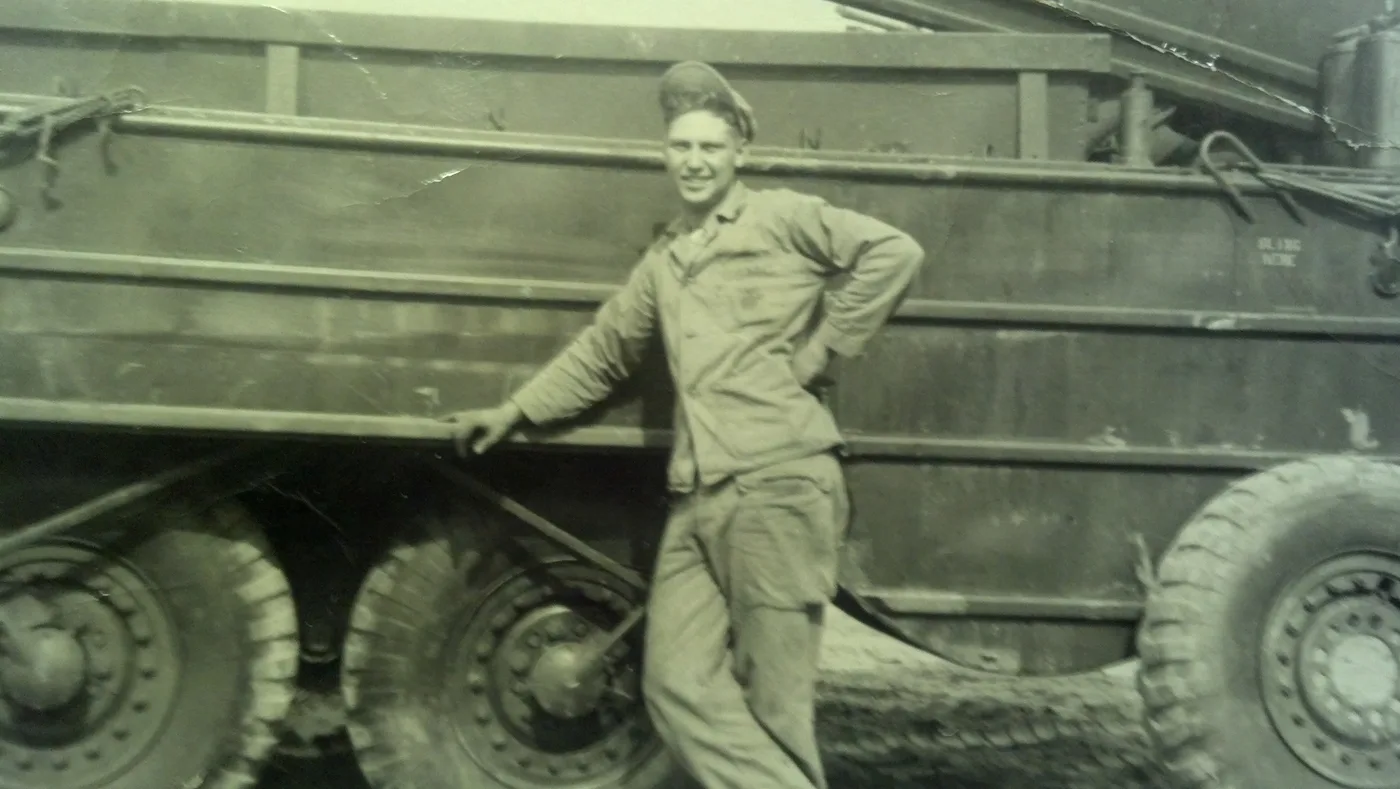 Sepia photograph of a smiling young man in a work uniform and cap, one hand on his hip, leaning against the side of a DUKW six-wheel-drive amphibious military vehicle stenciled SLING HERE.